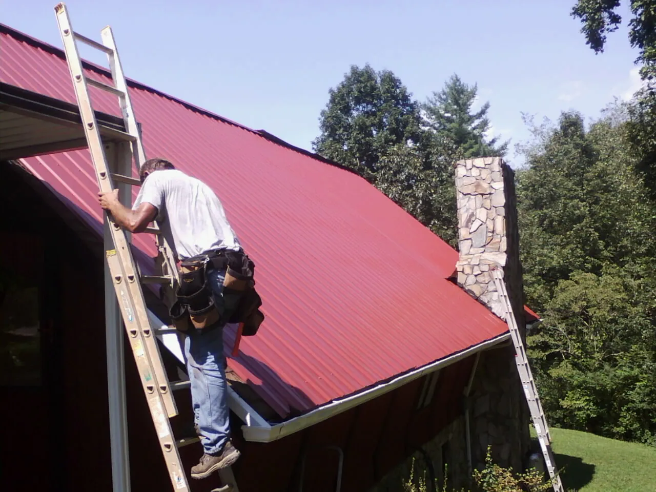 Roofer on a ladder beside a red metal roof and stone chimney — Soffit & Fascia Repair by Roofing Brothers in Tabiona